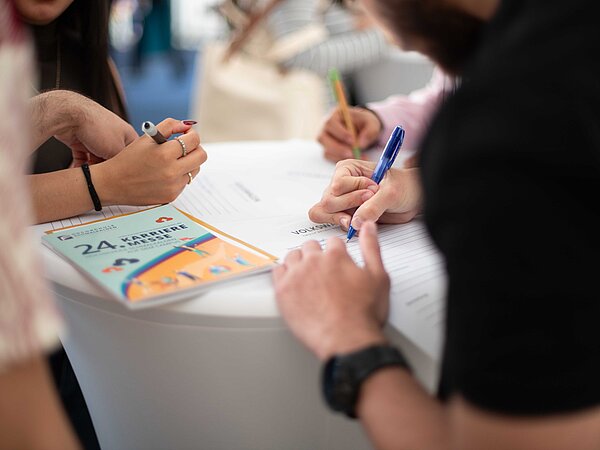 „Mehrere Personen füllen Formulare an einem weißen Tisch aus, mit Stiften und einem Heft mit der Aufschrift ‚24. KARRIERE MESSE‘ – Szene aus einer Messeanmeldung.
