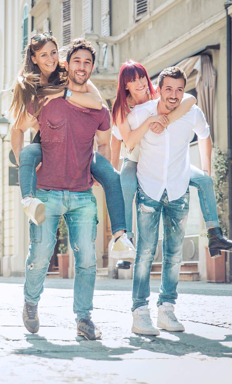 Das Bild zeigt sechs Freunde, die durch eine Stadt laufen. Jeder Mann trägt eine Frau auf dem Rücken. Sie alle lächeln und scheinen eine gute Zeit zu haben. Im Hintergrund sind Gebäude und Laternen zu sehen, die auf eine belebte Stadt hindeuten.