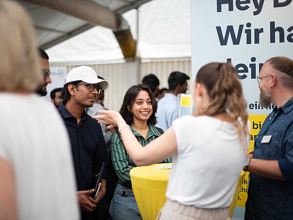 „Gruppe von Personen im Gespräch an einem gelben Tisch unter einem Zeltdach. Im Hintergrund ein Banner mit dem Schriftzug ‚Hey D… Wir hab… Leipzig‘, die Atmosphäre ist lebendig und offen.