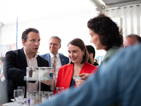 Personengruppe versammelt sich um einen Präsentationstisch in einem Messezelt. Ein Mann im dunklen Anzug erklärt etwas, während eine Frau in rotem Outfit aufmerksam zuhört. Auf dem Tisch stehen gestapelte Produktproben.
