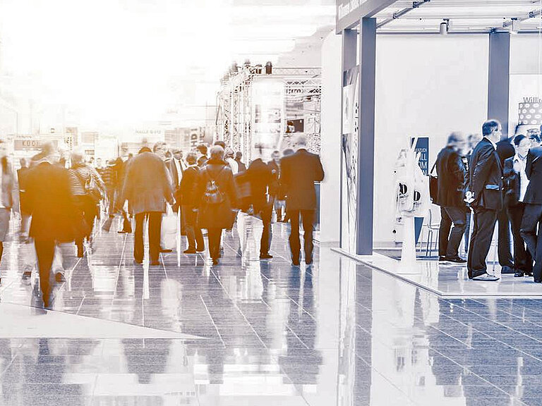 Menschen in Business- oder Semiformal-Kleidung bewegen sich durch eine helle Halle mit glänzendem Boden und Ausstellungsständen. Die Szene zeigt ein geschäftiges berufliches Event wie eine Messe oder Konferenz, geprägt von Interaktion und Networking.