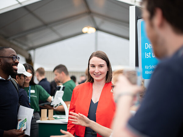 Frau im roten Blazer spricht mit Besuchern vor einem Bildschirm mit dem Text: 'Dein Studium ist unsere Mission – für Schüler:innen und Absolvent:innen' auf einer Karrieremesse.